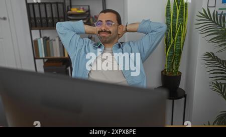 Young man with moustache wearing glasses relaxing in office chair in a modern workplace with plants and shelves in the background Stock Photo