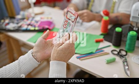 Two tailors work in an atelier, with new zealand dollars held foreground by a woman, implying a transaction or payment. Stock Photo