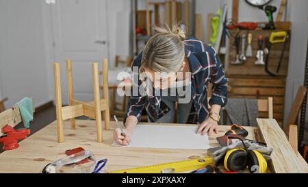 A young woman sketches a blueprint in a well-equipped carpentry workshop, surrounded by tools and wood. Stock Photo