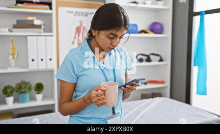 Indian woman healthcare professional on break in clinic room, holding mug and smartphone. Stock Photo