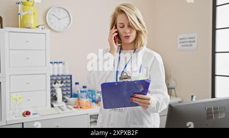 Blonde woman in lab coat talks on phone holding clipboard in laboratory Stock Photo