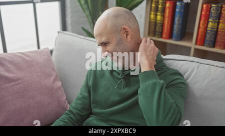 A bald man in a green hoodie smiles while scratching his head, sitting on a gray couch indoors with bookshelf background. Stock Photo