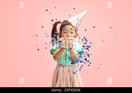 Waist up portrait of Sweet Asian Child Girl Blowing Blue Glitter to Camera While Standing Against Pink Isolated Background  Stock Photo