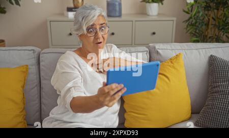 Elderly woman with grey hair and glasses sitting on a couch in a living room, looking surprised while holding a blue tablet Stock Photo