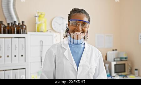 A smiling african american woman wearing goggles and a lab coat stands in a clinical laboratory setting. Stock Photo