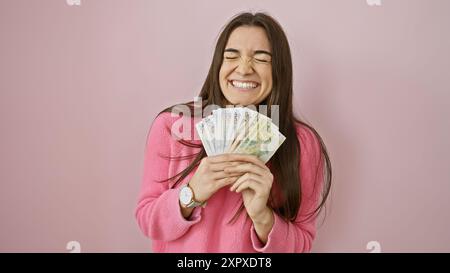 A cheerful young hispanic woman in a pink sweater joyfully holds peruvian currency before a pink wall. Stock Photo