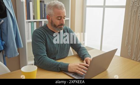 Mature man with grey hair and beard focused while using a laptop in a well-lit home interior. Stock Photo