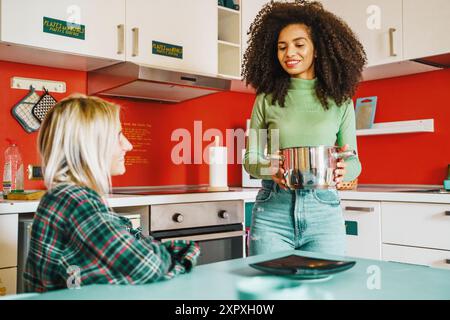Two young women, one with curly afro hair and one blonde, preparing food in a vibrant red kitchen. Showcases diversity, friendship, and modern lifesty Stock Photo