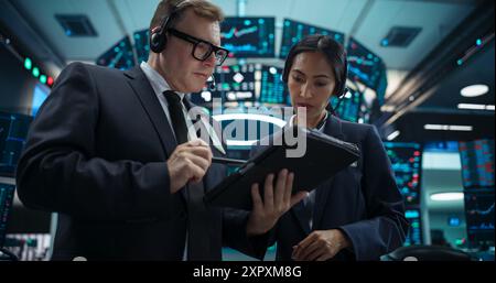Two Successful Stock Exchange Traders Using Tablet Computer, Discussing Commodities Investments for an International Business Partner, Working in an Office with Multiple Computer Screens Stock Photo