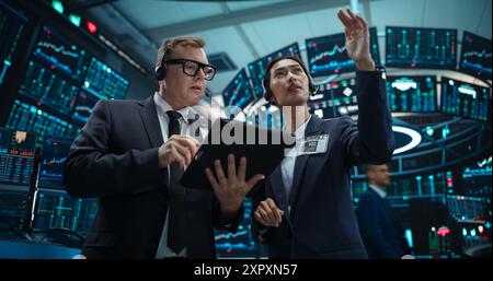 Two Successful Stock Exchange Traders Using Tablet Computer, Discussing Commodities Investments for an International Business Partner, Working in an Office Building with Multiple Computer Screens Stock Photo