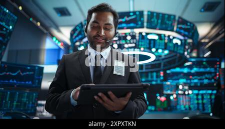 Portrait of a Young Indian Man Working in an International Stock Exchange Company: Happy Trader Using a Tablet Computer, Monitoring, Talking to a Corporate Business Partner in Headphones Stock Photo
