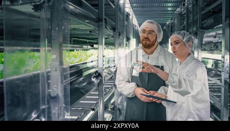 Couple Farming Experts Working at an Indoors Vertical Farm. Young Diverse Team of Gardening Specialists in Protective Contamination Uniform and Headwear Talking in a Corridor with Fresh Produce Stock Photo