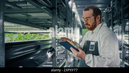 Portrait of a Biologist Analyzing Fresh Crops at Modern Vertical Farm Facility. Agricultural Engineer Working on a Tablet Computer, Improving and Optimizing Hydroponics System and Technologies Stock Photo