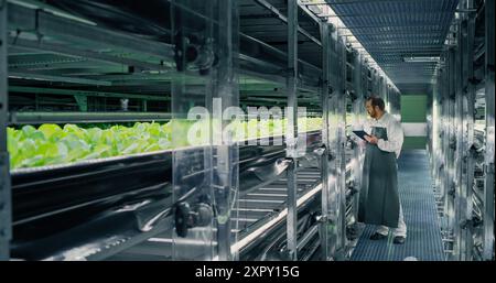 Agricultural Grower Working in a Corridor in a Vertical Farm Facility Next to Rack with Freshly Grown Plants. Hydroponics Technician Using Tablet Computer, Studying and Cultivating Crops Stock Photo