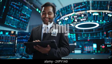 Portrait of a Young Indian Man Working in an International Stock Exchange Company: Happy Trader Using a Tablet Computer, Monitoring, Talking to a Corporate Business Partner in Headphones Stock Photo
