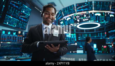 Portrait of a Young Indian Man Working in an International Stock Exchange Company: Happy Trader Using a Tablet Computer, Monitoring, Talking to a Corporate Business Partner in Headphones Stock Photo