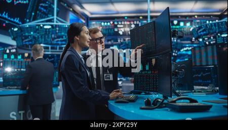 Asian Female and Male Stock Exchange Agents Using Desktop Computer, Discussing Investment Strategy for an International Business Client, Working in an Office Building Surrounded by Computer Screens Stock Photo