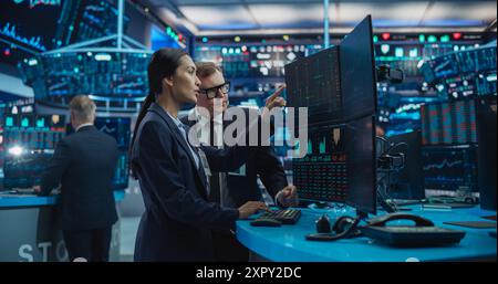 Asian Female and Male Stock Exchange Agents Using Computer, Discussing Investment Strategy for an International Business Client, Working in an Office Building Surrounded by Computer Screens Stock Photo