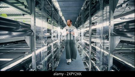 Biologist, Gardener, Scientist Walking Between Rows of Racks with Ecological Crops at a Vertical Farm. Horticulture Specialist Using Tablet Computer, Monitoring Growth and Nutrition Through a Software Stock Photo