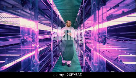 Farming Technician Walking Between Rows of Produce at Modern Vertical Farm With Neon Lights. Young Horticulturist in Protective Uniform Using Tablet Computer to Organize and Analyze Plant Information. Stock Photo