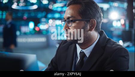 Portrait of a Young Indian Man Working in an International Stock Exchange Company: Happy Trader Sitting Behind a Desk with a Computer, Talking to a Corporate Business Partner in Headphones Stock Photo