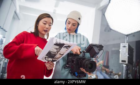 In A Modern Studio, An Asian Woman In A Red Sweater And A Caucasian Woman With Blue Hair, Both Young, Collaborate Over A Camera And Storyboard, Focusing On Creative Film Production And Advertising. Stock Photo