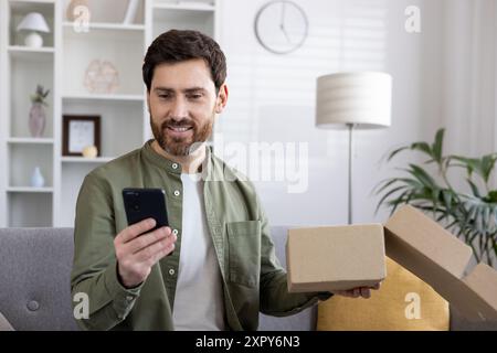 Smiling man using smartphone while unboxing package. Happy male checking phone. Concept of online shopping, delivery, e-commerce, and technology. Comfortable home environment. Stock Photo