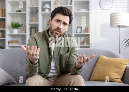 Man sitting on sofa expressing confusion during video call using phone in living room. Man making hand gestures looking at camera, feeling uncertain and perplexed. Stock Photo