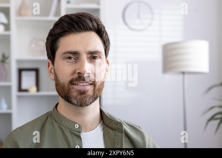 Smiling bearded man in casual clothes looking at camera in bright room. Positive expression and relaxed demeanor convey friendliness and approachability. Professional and casual mix for versatile uses. Stock Photo