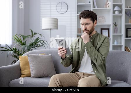 Man seated on sofa reading tablet at home. Thoughtful expression while using digital device. Modern living room background. Concept of technology, leisure, home office, remote work, reading, phone Stock Photo