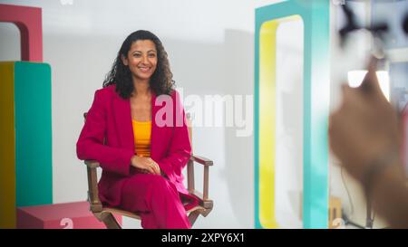 Young Middle Eastern Female Host In Vibrant Pink Blazer And Yellow Top, Seated On Director's Chair On Colorful Film Set, Smiling At Camera, With Blurred Videographer In Foreground. Stock Photo