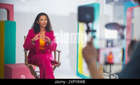 Vibrant Scene Captures A Black Female Host In A Pink Suit, Engaging Animatedly In A Colorful Studio Setup, Surrounded By Modern Geometric Props, During A Lively Video Recording Session For A Talk Show Stock Photo