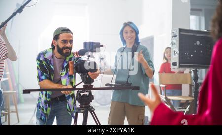 Young Caucasian Male And Female Filmmakers Collaborate On A Film Set, Adjusting Camera Equipment With Enthusiasm, Surrounded By Diverse Crew Members In A Bright, Modern Studio Environment. Stock Photo