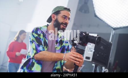 Young Male Cinematographer With Beard, Wearing A Colorful Jacket And Cap, Operates A Camera In A Studio Setting, With A Female Assistant In Background, Focusing On A Creative Shoot. Stock Photo