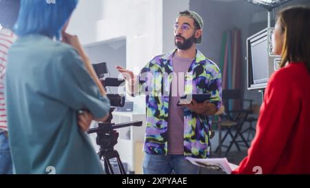 Young Caucasian Male Director With Beard And Glasses, Instructing Diverse Team On Film Set, Holding Tablet, Surrounded By Camera Equipment And Crew In Creative Studio Environment. Stock Photo