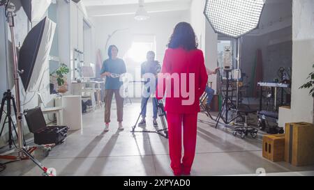 Young Diverse Team Of Filmmakers In A Bright Studio Setting, With A Woman In A Red Suit Directing The Shoot, Surrounded By Cameras, Lights, And Crew Members Focused On Creating Engaging Content. Stock Photo