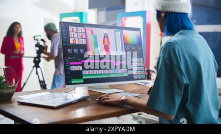 Young Caucasian Female Editor With Blue Hair Works On Video Editing Software, While A Hispanic Male Videographer Records A Black Female Host In A Vibrant Studio Setting, Enhancing Media Production. Stock Photo