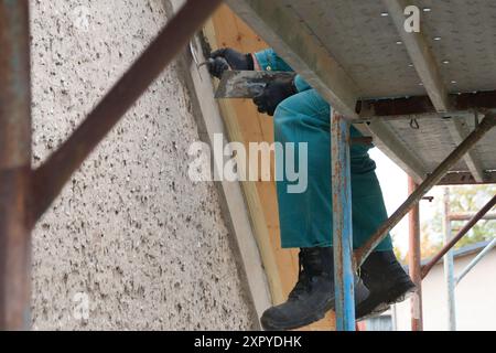 Close-up of the legs of a construction worker sitting on a scaffold wearing safety shoes and repairing something on the roof gable with trowels and ce Stock Photo