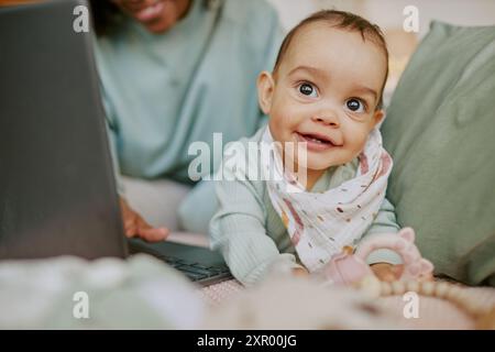 Baby boy wearing flax bib fooling around on comfortable bed while his mother working remotely via laptop in background Stock Photo