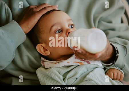 Medium close up of male infant wearing bib and eating formula from baby bottle while caring hand of his mother patting his head Stock Photo