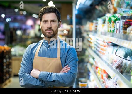 Male grocery store worker standing confidently in store aisle with arms crossed, wearing apron and blue shirt. Shelves stocked with various products visible in background, suggesting retail, business Stock Photo