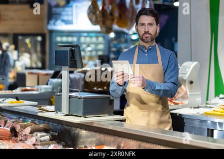 Male butcher in apron using tablet behind counter at deli shop. Display of various meats and sausages visible. Digital transformation in traditional retail setting Stock Photo