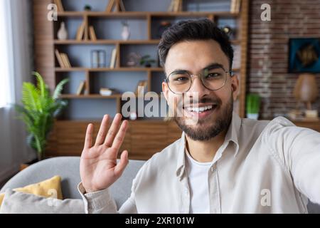 Young man waving and smiling during video call using phone at home. Casual and friendly setting with bookshelf in background. Concept of remote work, communication, and connection. Stock Photo