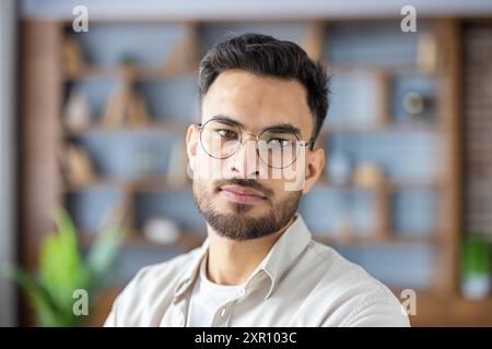 Confident young man with glasses looking at camera in modern office setting. Professional appearance, stylish eyeglasses, relaxed expression. Represents business, confidence, and modern work environment. Stock Photo