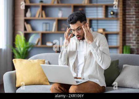 Shocked man sitting on couch with laptop on lap, hands on head, experiencing surprise, stress, or frustration. Working from home, casual wear, modern living room, using smartphone Stock Photo