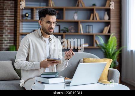 Young man studies remotely from home using laptop for video call with mentor, taking notes and discussing. Concept includes online education, remote learning, and modern technology. Stock Photo