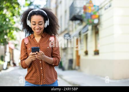 Cheerful young woman wearing headphones and using phone while walking outdoors. Enjoying music, smiling, and staying connected. Vibrant city background with casual attire and positive vibes. Stock Photo
