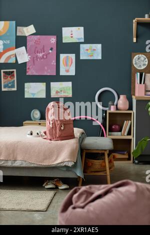 Cozy kids bedroom with a study area featuring a backpack on gray sofa, posters displaying fun artwork on dark wall, and neatly arranged storage for a tidy organized space Stock Photo