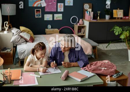 Grandmother sitting with granddaughter and assisting with schoolwork at desk in cozy bedroom with educational posters and art supplies on the table Stock Photo