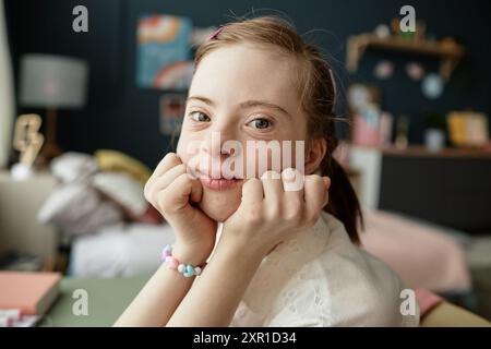 Portrait of young girl with Down syndrome resting chin on hands and smiling. Colorful room in background with artwork and toys providing vibrant atmosphere Stock Photo